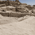 A high-angle view looking down at the massive Unfinished Obelisk carved directly into the pink granite bedrock of an ancient quarry in Aswan, under a clear sky. Caption A fascinating glimpse into ancient craftsmanship: the monumental Unfinished Obelisk