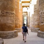 A Top Class Egypt Travel client, wearing a hat and carrying gear, walks between the massive, hieroglyph-carved columns of the Great Hypostyle Hall at Karnak Temple during a private, expert-led tour in Luxor