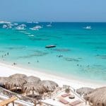 A wide panoramic daytime view of a white sand beach in Hurghada lined with thatched umbrellas, looking out over clear turquoise Red Sea waters dotted with anchored boats.
