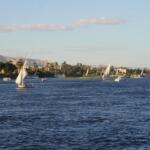 Traditional Egyptian felucca sailboats sailing on the Nile River in Luxor with lush riverbanks and desert hills in the background.9 Days Cairo and Nile cruise.