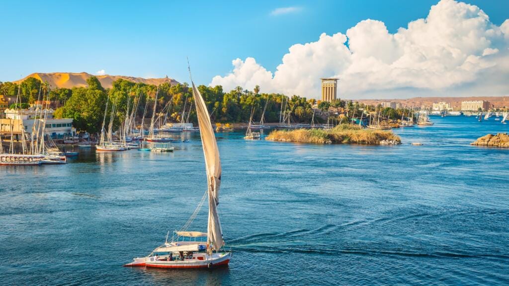 A traditional Egyptian felucca sailboat with a large white sail gliding gracefully on the blue Nile River near Aswan, surrounded by lush riverbanks and a clear sky