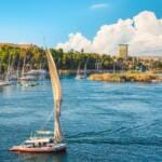 A traditional Egyptian felucca sailboat with a large white sail gliding gracefully on the blue Nile River near Aswan, surrounded by lush riverbanks and a clear sky