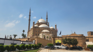 A low-angle view looking up at the monumental Muhammad Ali Mosque within the Citadel of Salah El-Din in Cairo, showcasing its grand stone domes, twin pencil minarets, and historic fortified walls against a blue sky.