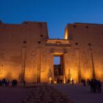 A high-angle perspective of the massive stone pylon entrance of the Temple of Horus at Edfu, framed by a paved courtyard and columns under a dramatic orange sunset sky.