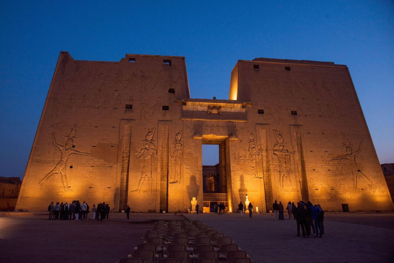 A high-angle perspective of the massive stone pylon entrance of the Temple of Horus at Edfu, framed by a paved courtyard and columns under a dramatic orange sunset sky.