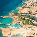 A high-angle aerial view of the Red Sea coastline in Hurghada, showcasing deep blue waters shifting to a turquoise lagoon beach and a premium resort hotel layout along the coast.