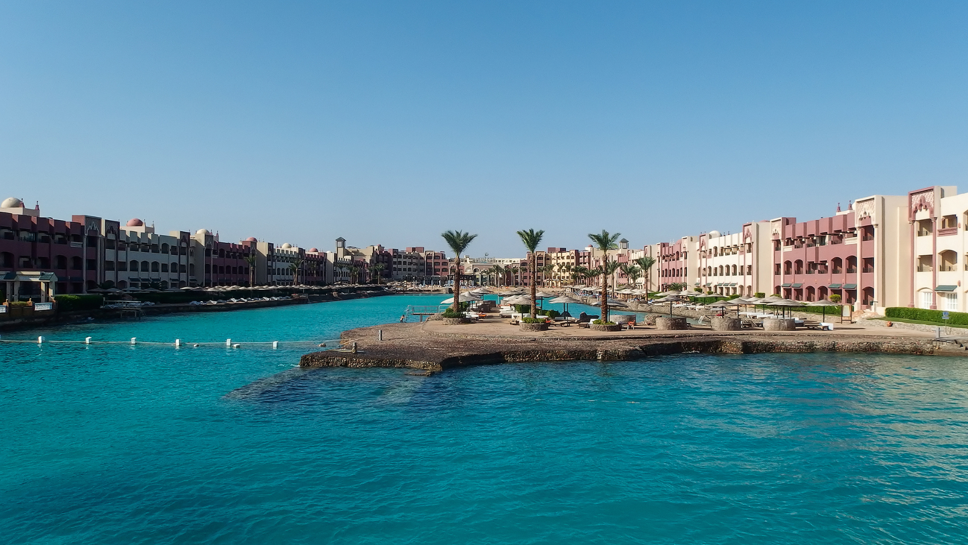 A scenic daytime view of a luxury resort in Hurghada featuring vibrant turquoise sea water in a coastal lagoon or marina channel, flanked by multi-story resort buildings and small palm trees under a clear blue sky.