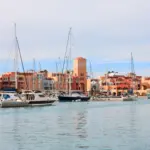 A scenic daytime view of El Gouna Marina featuring luxury yachts and sailboats moored in the calm harbor waters, with Mediterranean-style terracotta and pastel resort buildings lining the waterfront.