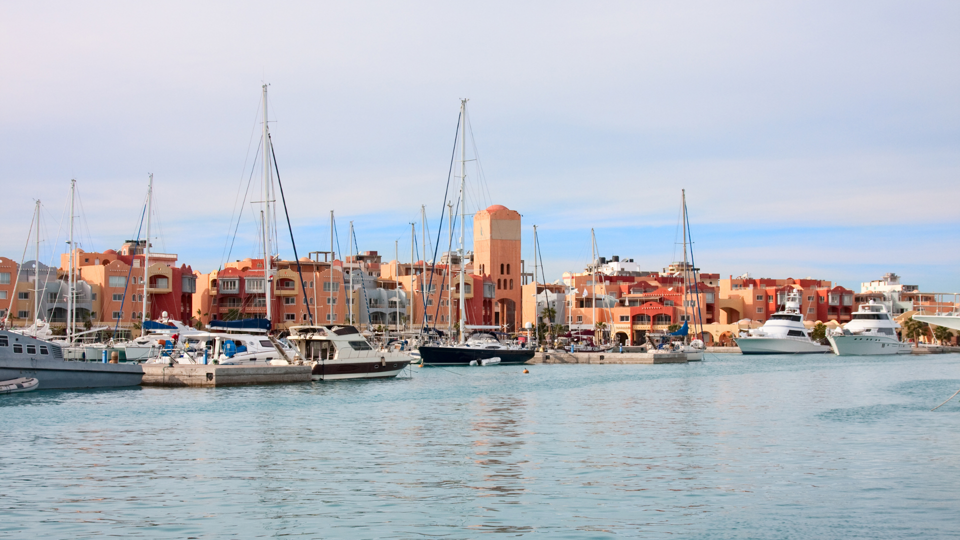 A scenic daytime view of El Gouna Marina featuring luxury yachts and sailboats moored in the calm harbor waters, with Mediterranean-style terracotta and pastel resort buildings lining the waterfront.