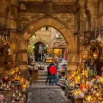 A vibrant, eye-level shot looking through a historic stone archway into a bustling alley at Khan El-Khalili bazaar, crowded with market stalls displaying glowing brass lanterns and traditional Egyptian handicrafts.