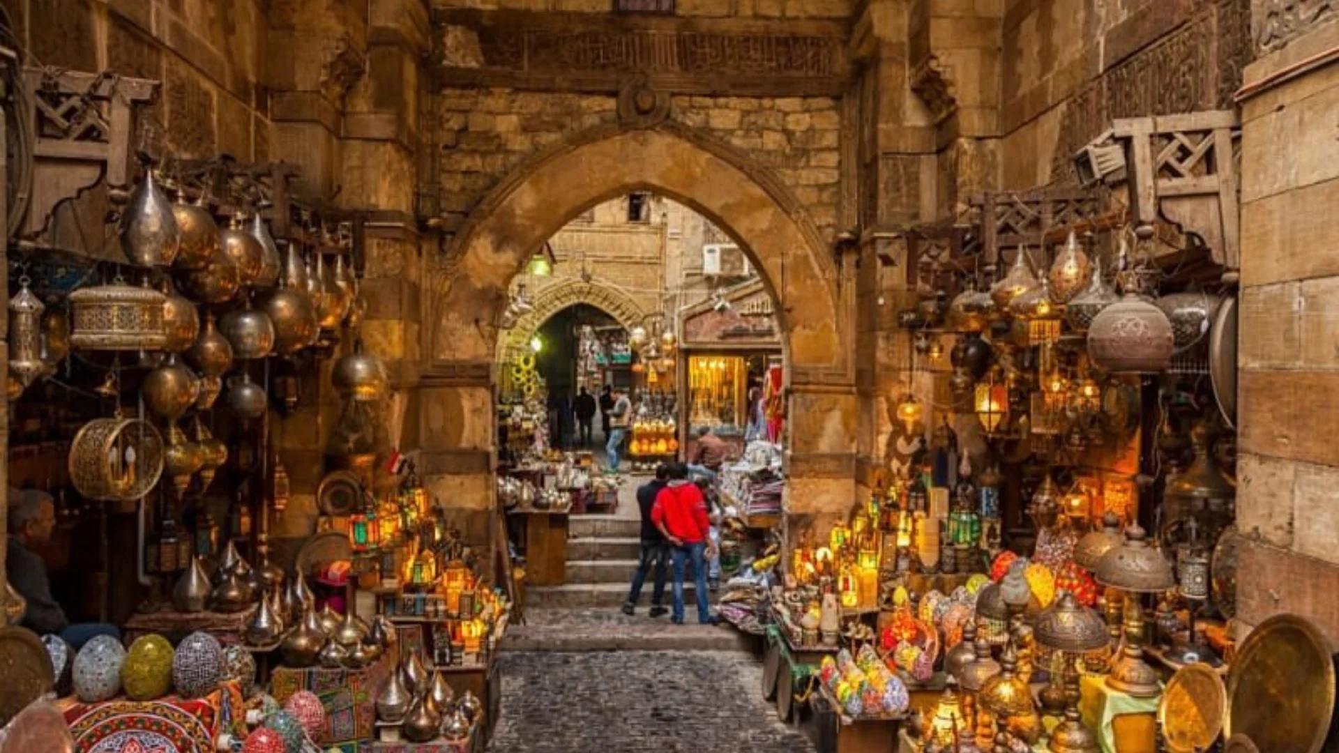 A vibrant, eye-level shot looking through a historic stone archway into a bustling alley at Khan El-Khalili bazaar, crowded with market stalls displaying glowing brass lanterns and traditional Egyptian handicrafts.