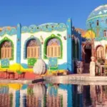 A view from the Nile River of a vibrant Nubian village in Aswan, showcasing traditional brightly painted blue and white houses along the rocky shoreline.