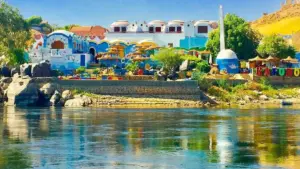 A view from the Nile River of a vibrant Nubian village in Aswan, showcasing traditional brightly painted blue and white houses along the rocky shoreline.