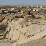 A high-angle view looking down at the massive Unfinished Obelisk carved directly into the pink granite bedrock of an ancient quarry in Aswan, under a clear sky. Caption A fascinating glimpse into ancient craftsmanship: the monumental Unfinished Obelisk