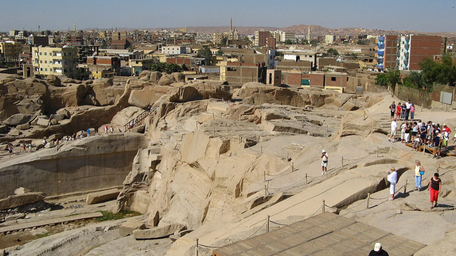 A high-angle view looking down at the massive Unfinished Obelisk carved directly into the pink granite bedrock of an ancient quarry in Aswan, under a clear sky. Caption A fascinating glimpse into ancient craftsmanship: the monumental Unfinished Obelisk