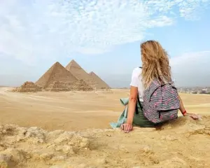 A female traveler with a backpack sitting in the desert sand, looking at the Great Pyramids of Giza during a day tour.