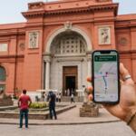 A first-person perspective holding a smartphone with a GPS map app in front of the iconic red facade of the Egyptian Museum in Tahrir Square.