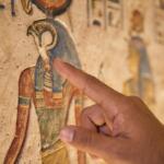 A close-up shot of a professional guide's hand pointing toward detailed ancient hieroglyphics and a depiction of the god Horus on a limestone temple wall.