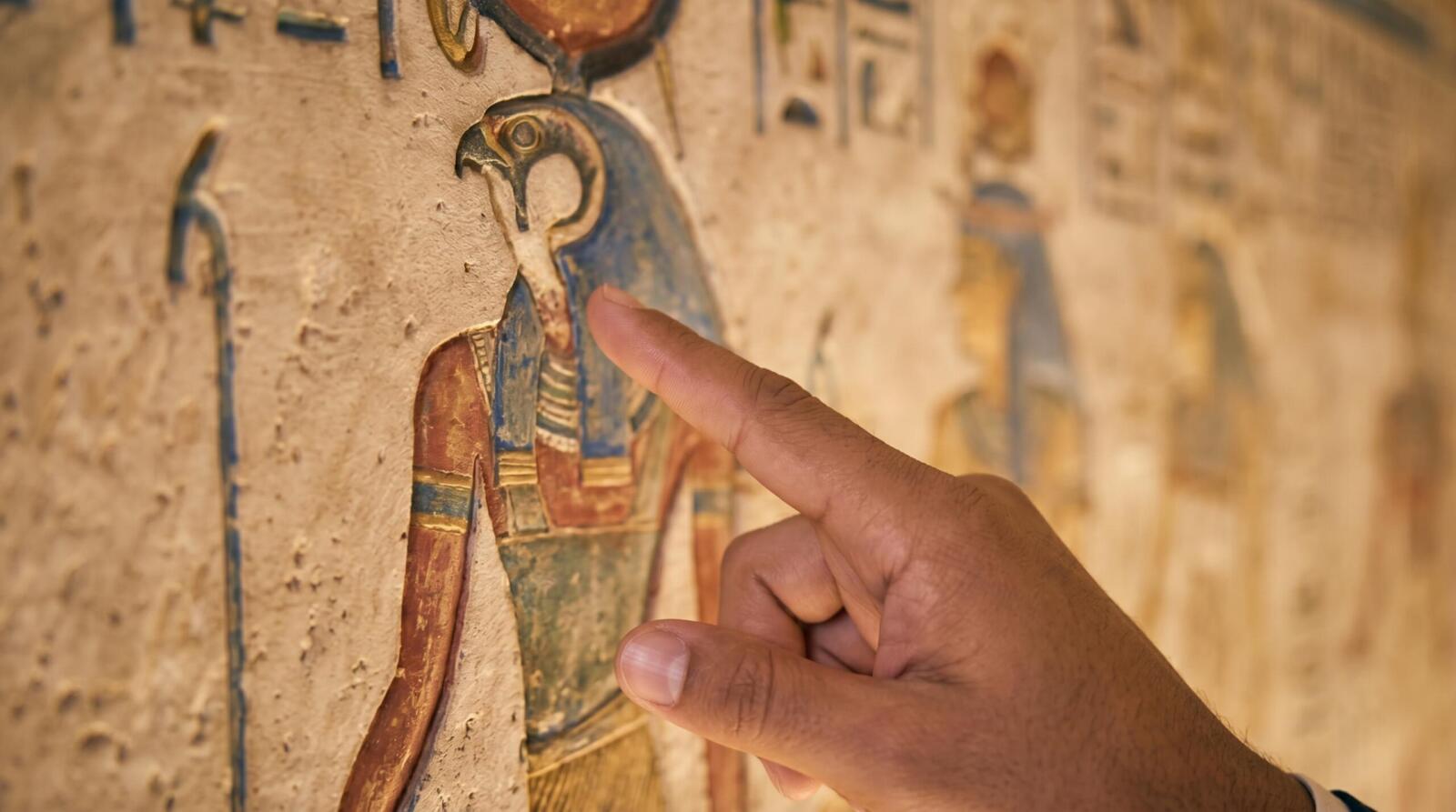 A close-up shot of a professional guide's hand pointing toward detailed ancient hieroglyphics and a depiction of the god Horus on a limestone temple wall.