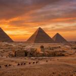 Wide-angle panoramic view of the three Great Pyramids of Giza and the Sphinx under a vibrant orange and purple sunset sky.