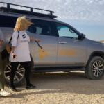 Two women standing next to a silver 4x4 safari SUV with the Gazelle Crossover logo in a sunny desert landscape.
