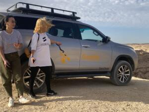 Two women standing next to a silver 4x4 safari SUV with the Gazelle Crossover logo in a sunny desert landscape.