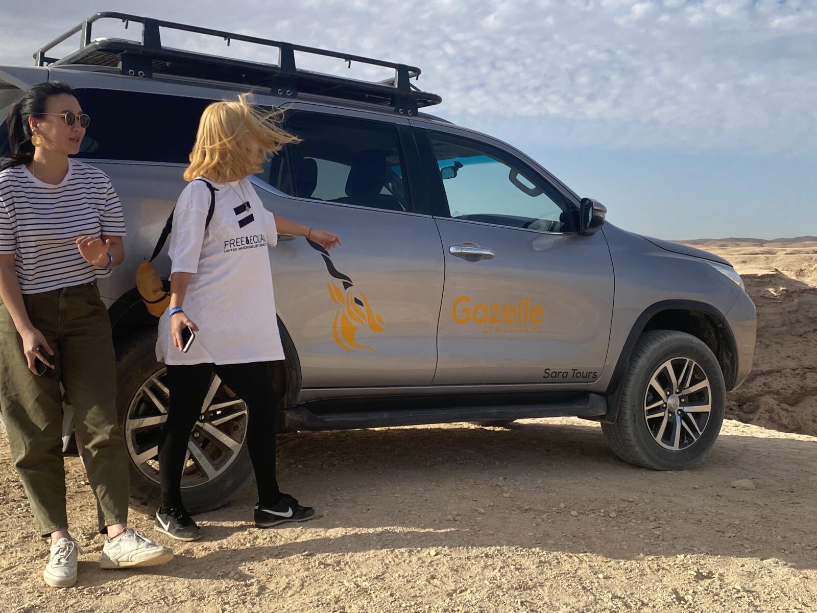 Two women standing next to a silver 4x4 safari SUV with the Gazelle Crossover logo in a sunny desert landscape.