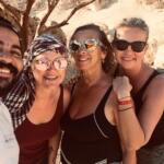 A group of four smiling travelers, including a man and three women wearing sunglasses, posing for a selfie during a desert safari trip.