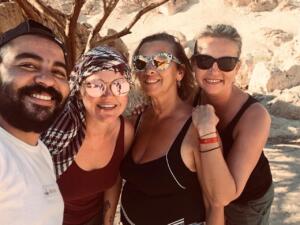 A group of four smiling travelers, including a man and three women wearing sunglasses, posing for a selfie during a desert safari trip.