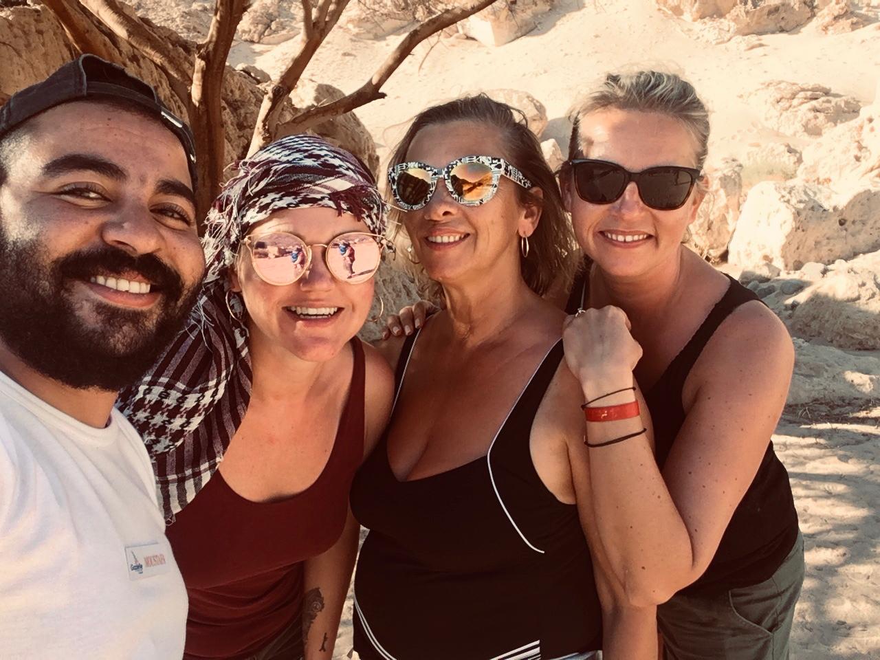 A group of four smiling travelers, including a man and three women wearing sunglasses, posing for a selfie during a desert safari trip.