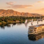 A 5-star luxury Nile cruise ship sailing at sunset with white-sailed feluccas and desert mountains.