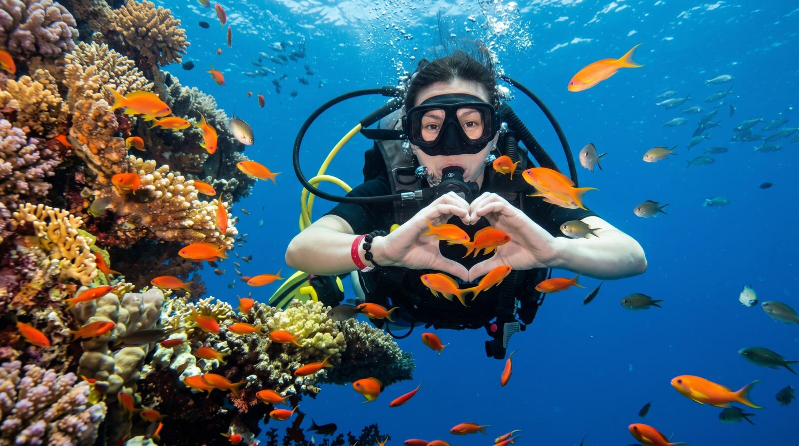 A scuba diver underwater making a heart shape with their hands during a luxury dive in Hurghada, surrounded by a vibrant coral reef and bright orange fish.