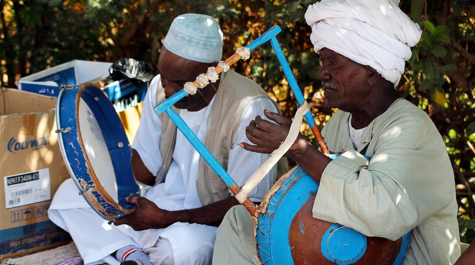 A close-up view of a Nubian woman wearing a black patterned headscarf singing alongside male musicians playing frame drums in white turbans.