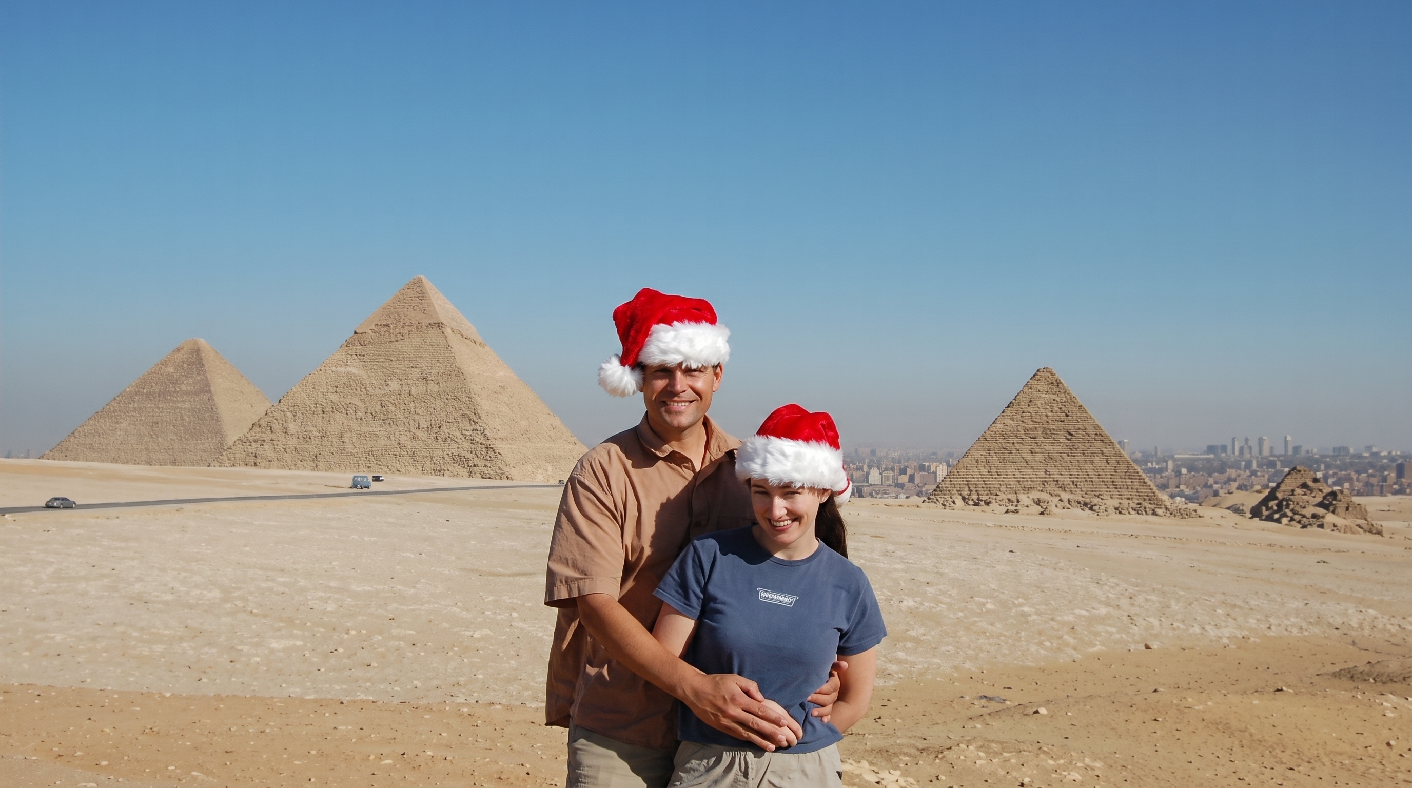 A smiling couple wearing Santa hats hugging in front of the spectacular Giza Pyramids during a sunny winter day